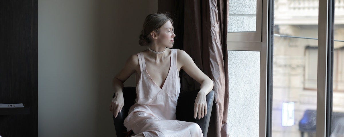 A beautiful woman with her hair up, wearing a pale pink shift dress, red heels, and a diamante choker. She is sitting in a chair in a hotel room, looking out of the window at the street below.