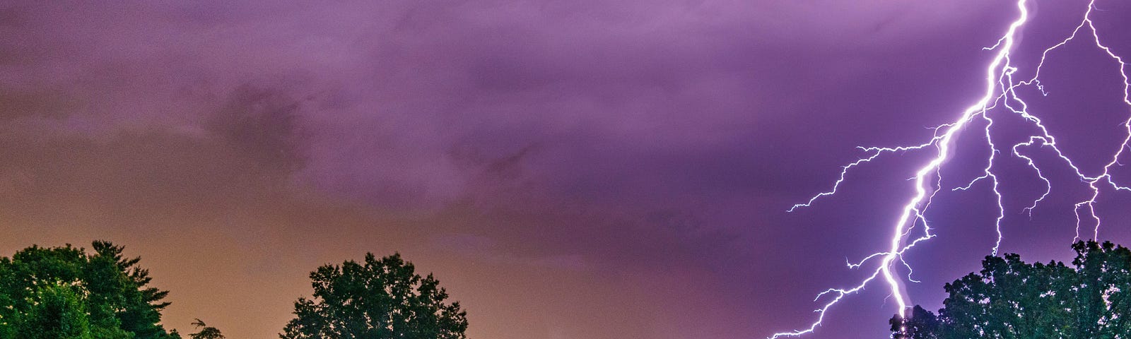 color photograph of brilliant lightning striking in the violet sky with a green grassy foreground and a small white house in the left front corner
