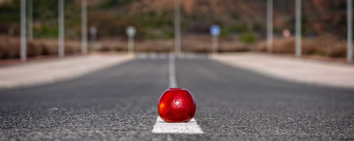 A shiny red apple lying on the white meridian line of an otherwise deserted road.