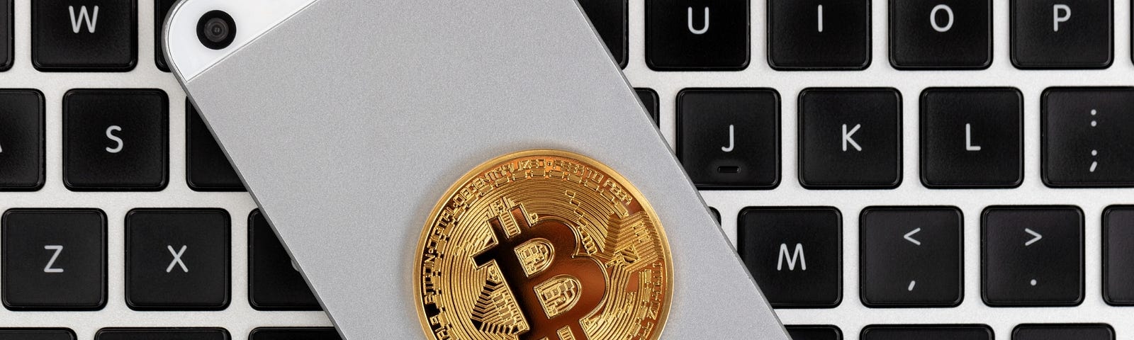 A Bitcoin laying on the back of a cell phone which is on top of a Macbook keyboard