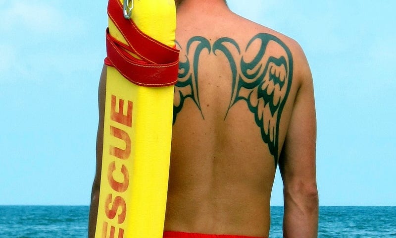 Lifeguard, with angel wings tattoo, standing on the beach holding rescue float over left shoulder, looking out over the water.