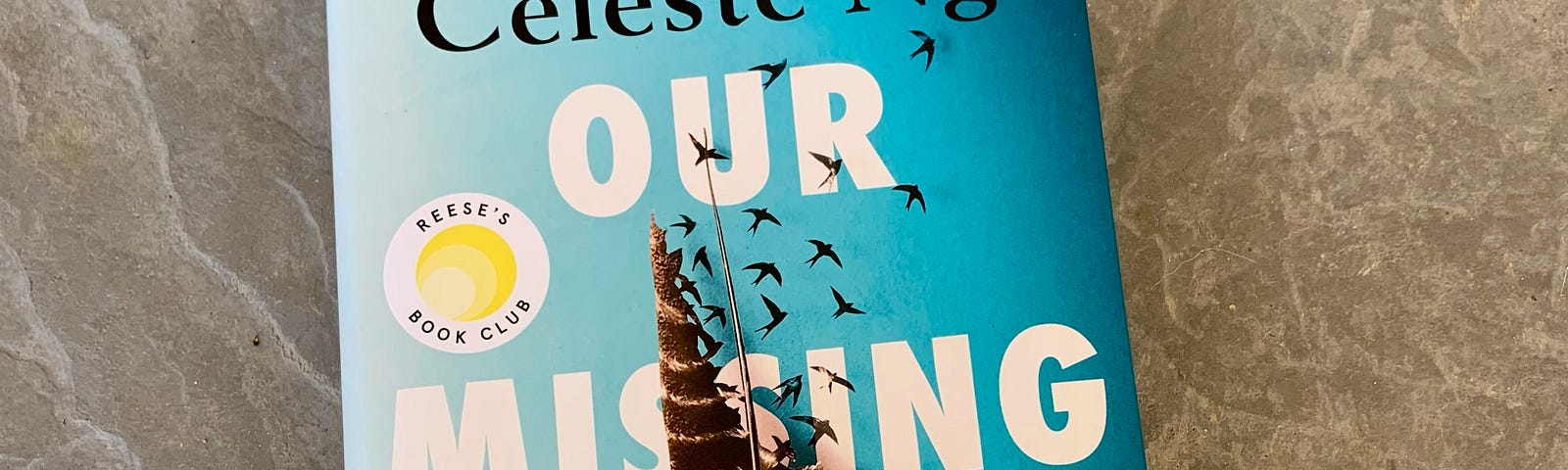 A hardcover copy of Our Missing Hearts by Celeste Ng lays on a gray concrete slab. The book has a blue background with a gray and white striped feather. The feather has part of it missing as it turns into birds flying up and away.