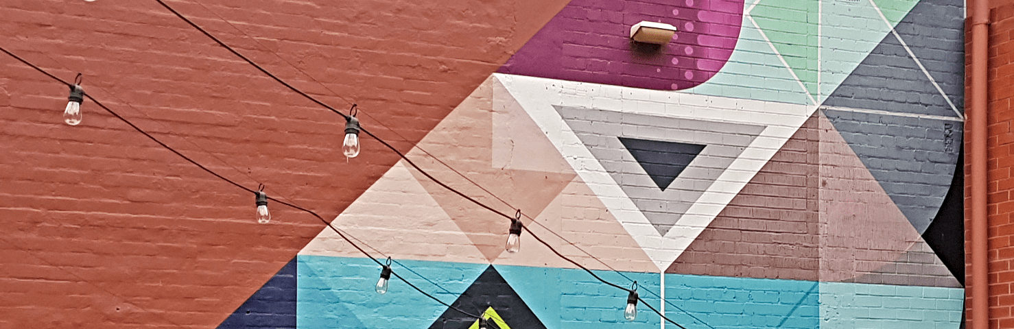 A brick wall is painted in patterned geometric shapes in purples, greens, yellows, browns, orange, rust-red, white, black, greys and shades of blue. Two cords of lightbulbs are strung diagonally across the wall. A painted down-pipe borders the photo’s right frame.