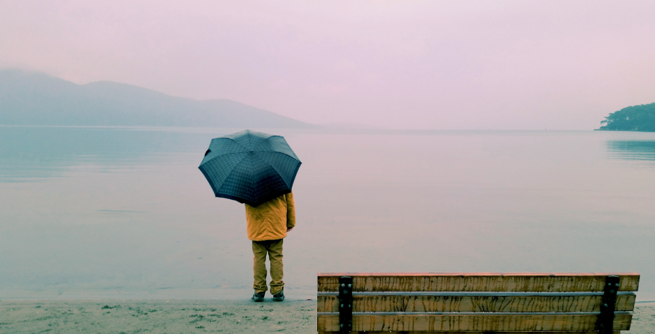 A person holding an umbrella looking at water with an empty bench near them