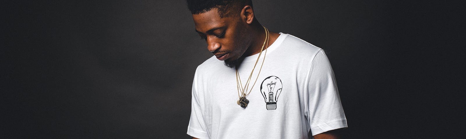 Black man in white t-shirt looking down against a dark background.