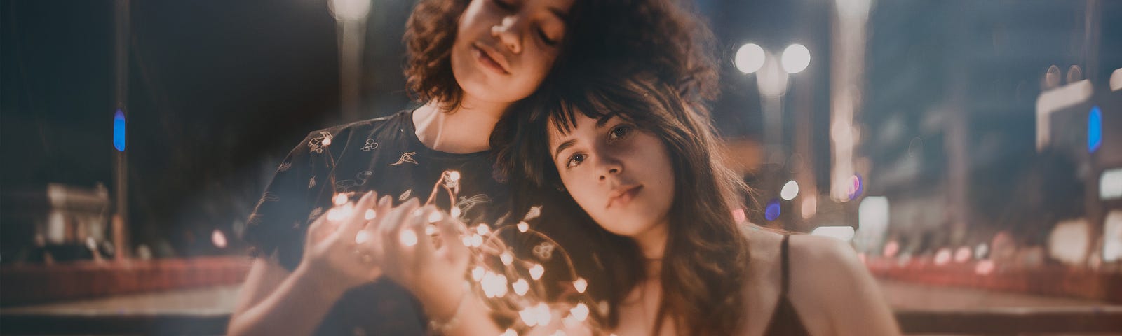 Two young women leaning against each other with a string of fairy lights between them