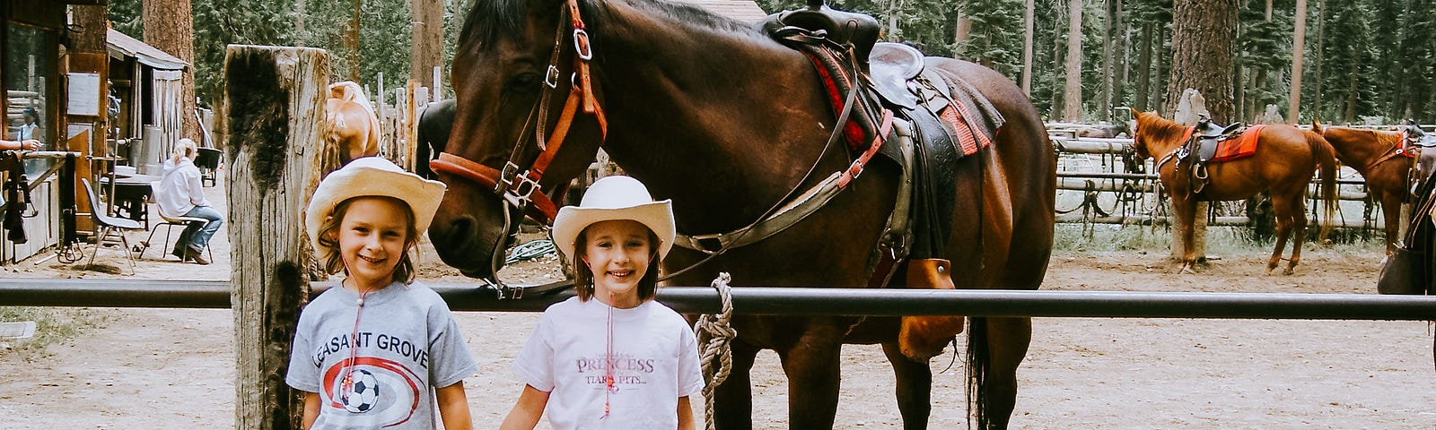 Emma and Maddie hold hands posing in front of a horse at a ranch surrounded by trees.