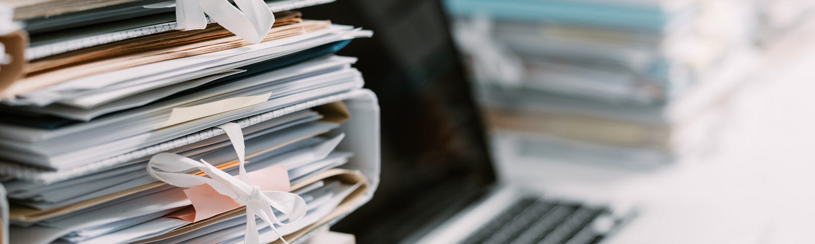 Photograph of a desk, with a pile of papers and folders in the foreground. A laptop with blank screen is behind them.