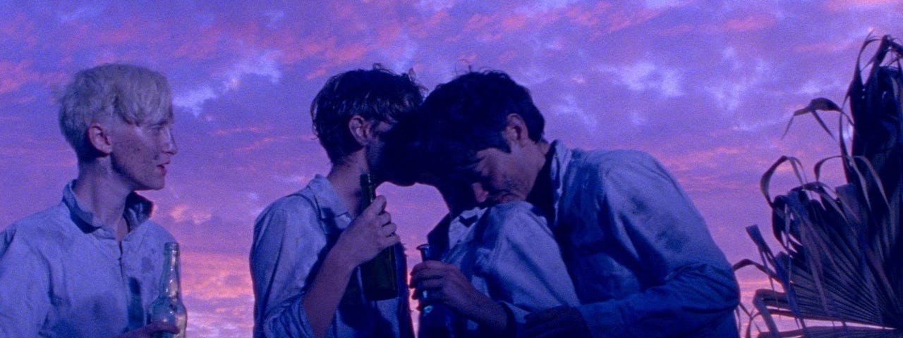 Violet pictures of the Wild Boys over a clear sky huggging one another on what seems to be a beach.