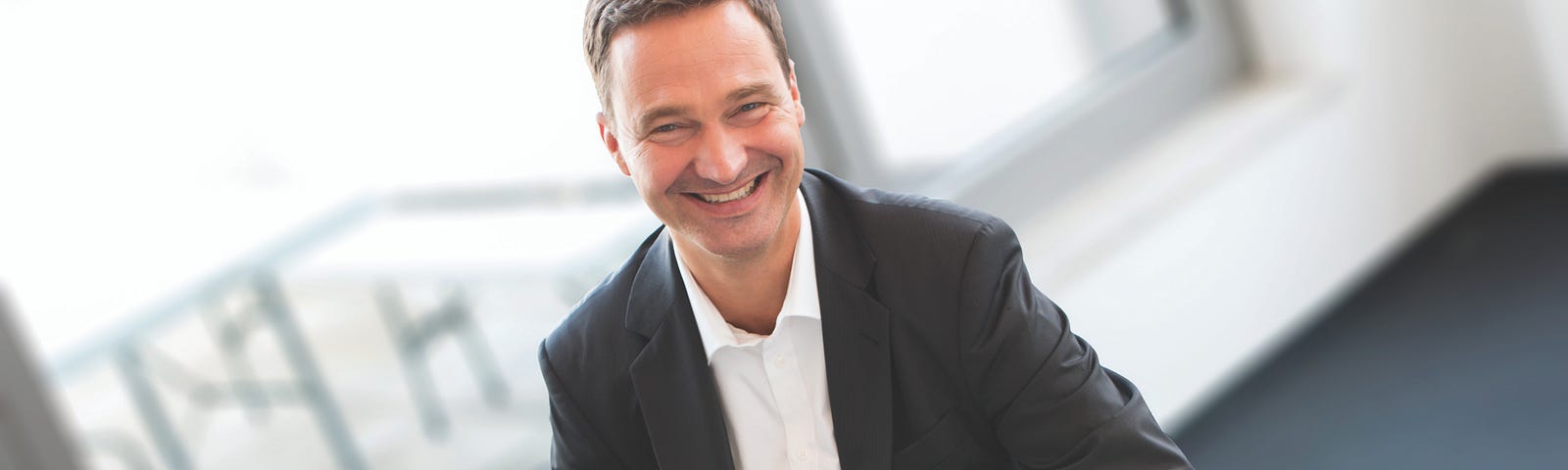 Alex von Frankenburg wears a black suit and smiles, sitting in an office with wide windows on a sunny day