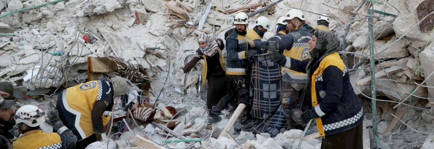 A group of people wearing yellow vests showing they are part of the White Helmets work on top and in front of rubble. The men wear white helmets. Several women wear head coverings.