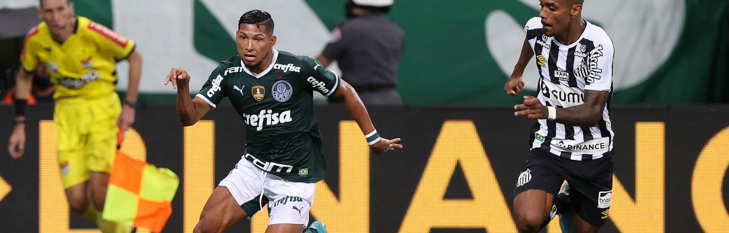 Rony corre e enfrenta a marcação de jogador Lucas Pires, do Santos. Os dois são negros, de altura mediana. Rony veste o uniforme tradicional do Palmeiras, camisas e meias verdes e calção branco, e conduz a bola de pé direito; Lucas Pires usa a camisa listrada do Santos, calção e meias pretas, e observa Rony a cerca de um metro de distância.