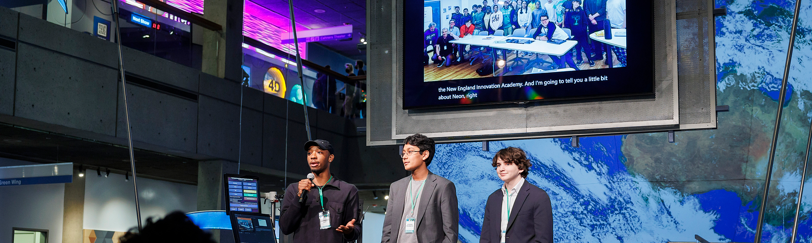 Three students stand on a stage with a large monitor behind them displaying a group of people and the words “Thank you.”