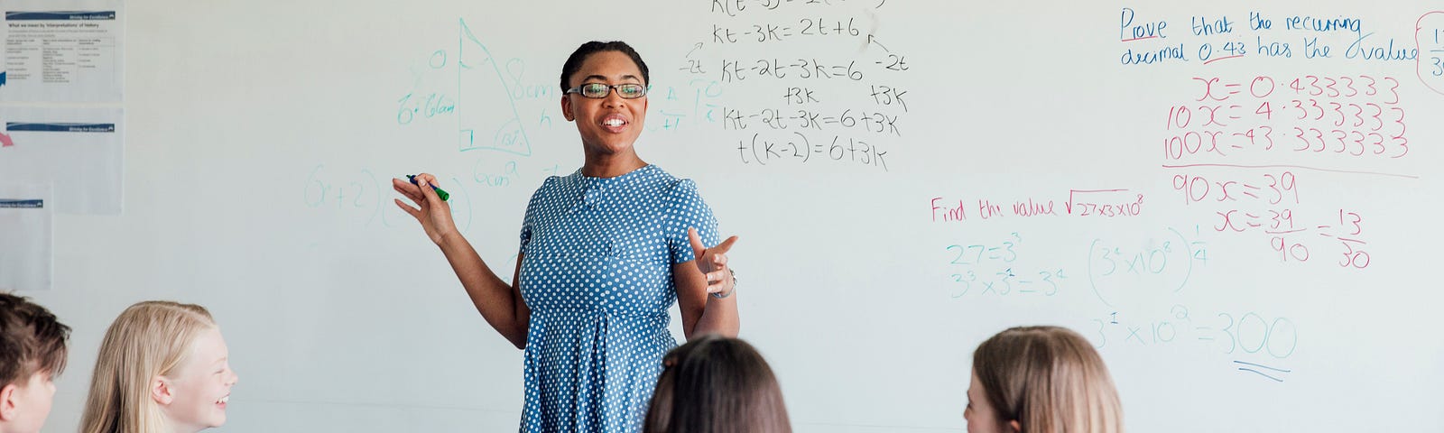 Woman in a light blue polka-dot dress, standing in front of a white board with math equations while 3 female students in school uniforms look forward at her.