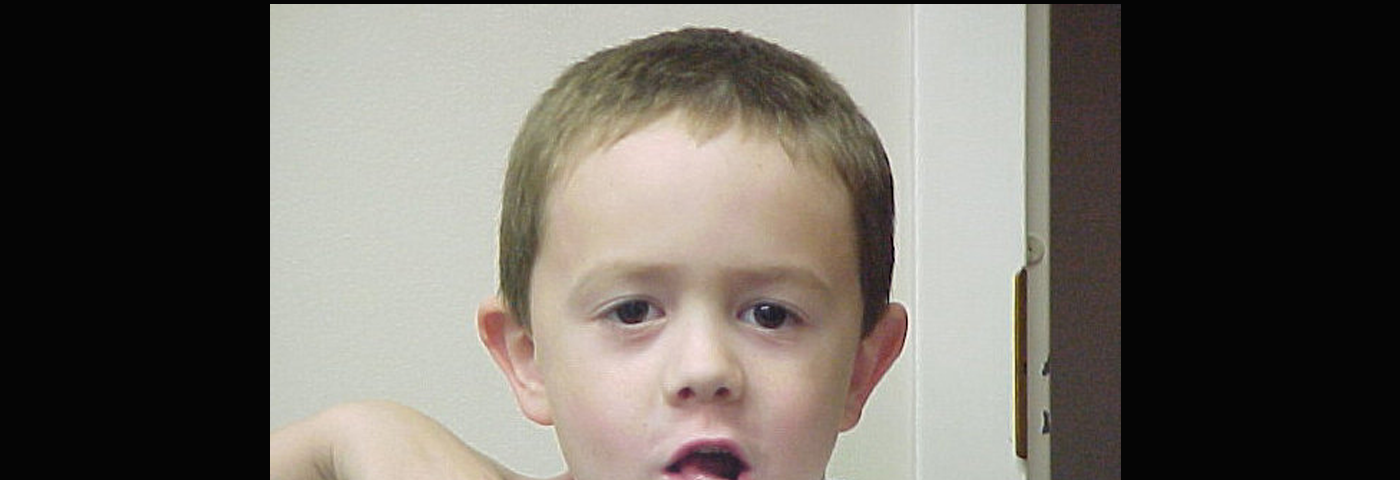 A young boy making a silly face. He is Caucasian with light brown hair in front of a white wall. His right hand is above his right shoulder and he is pointing downward toward the camera.