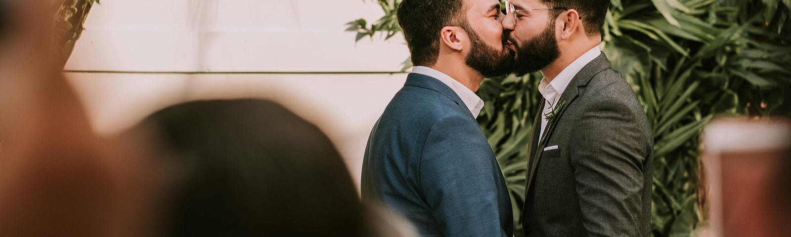A photo shows two grooms dressed in suits kissing. The grooms are in focus, while the wedding guests who are looking on are shown out of focus and from behind. Behind the grooms is some tropical foliage.