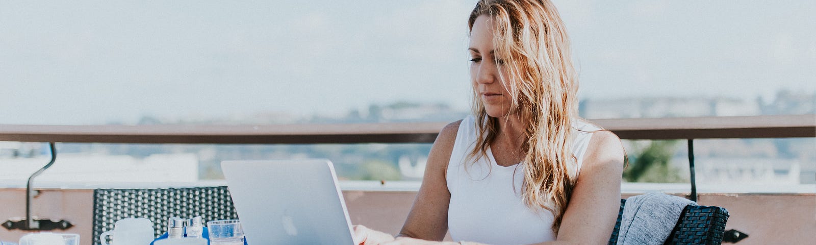 Image of a woman working on her laptop at a table on a balcony overlooking a cityscape