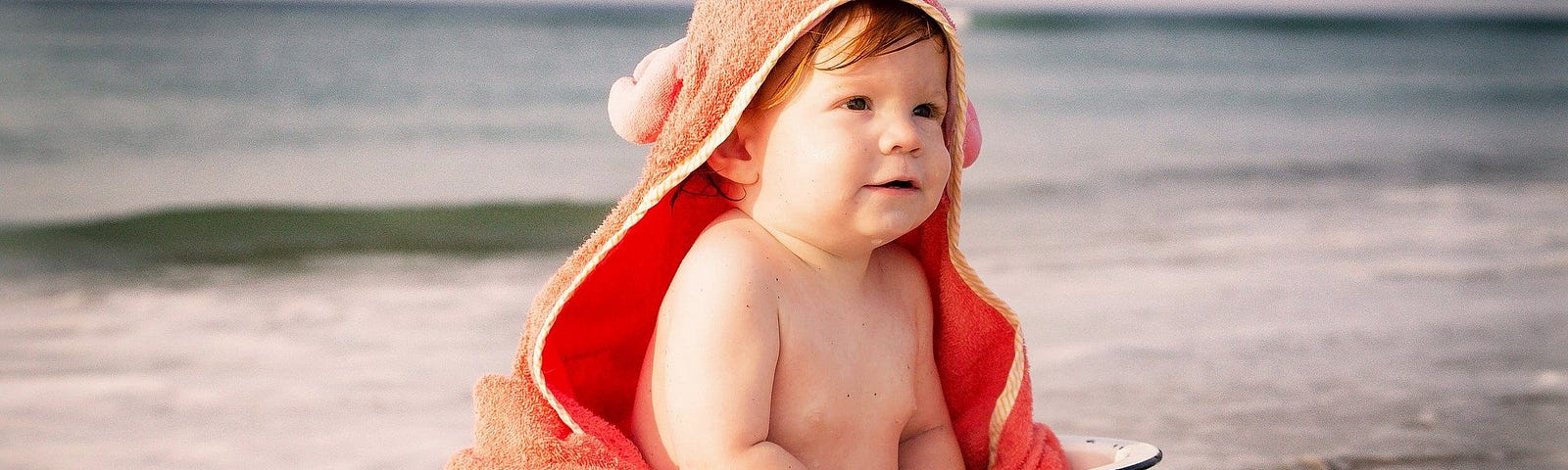 Baby wearing crab towel sitting in an old-fashioned hand basin at the beach