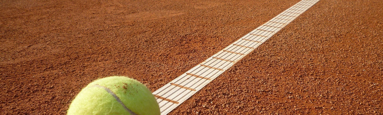 Brown ground of a tennis court with a white boundary line. A yellow tennis ball sits on the white boundary line.