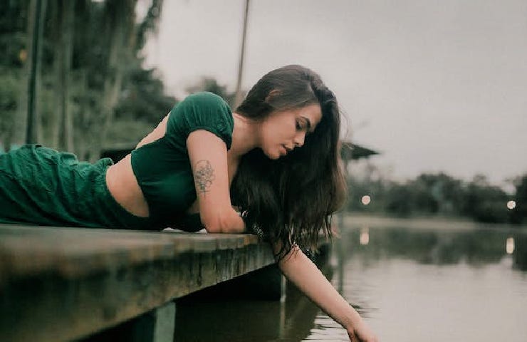 A woman dressed in green lays on a dock while having her hand in the water below.