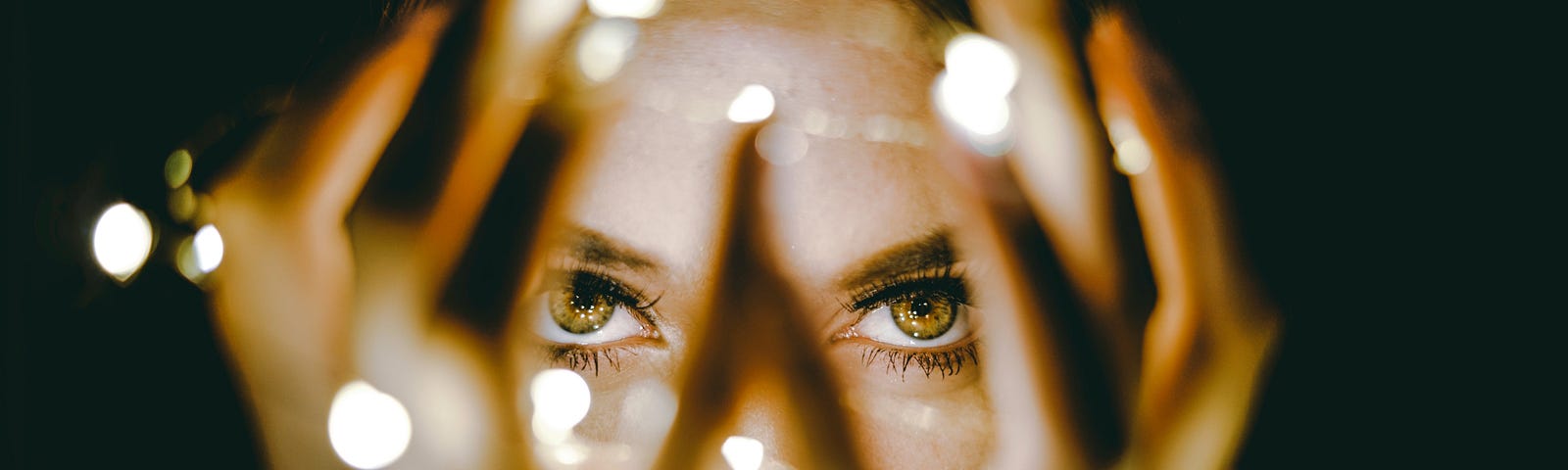 Woman with her hands open around her face with fairy lights wrapped around her hands. Focus is on her hazel eyes.