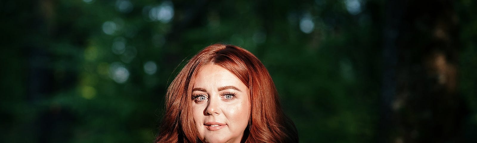 Close-up of attractive, vibrant woman in dusky pink blouse and long thick locks of red hair, standing in a shaded forest. Spotlight is on her face. By photographer Lisa McConnellogue. Ciara McCarron from Derry, Northern Ireland, is a Trauma-Informed Coach, Mentor & Therapist, Hypnotherapist and Founder of Redesign Your Vibe.