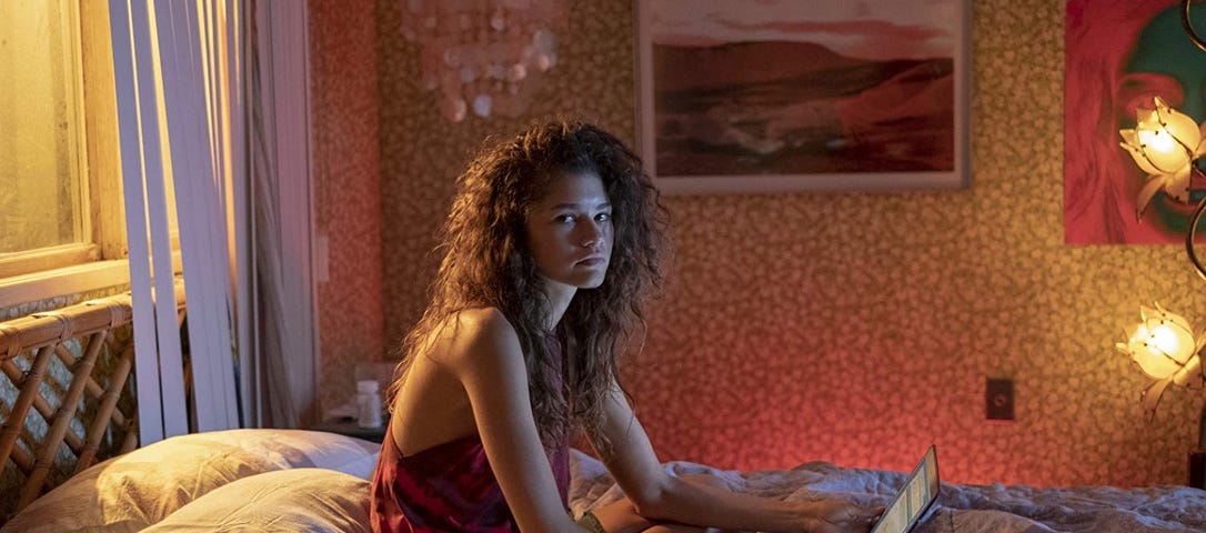 rue, personagem de Zendaya, está sentada na cama com seu laptop em sua frente. A atriz olha o fotógrafo a sua direita de pé
