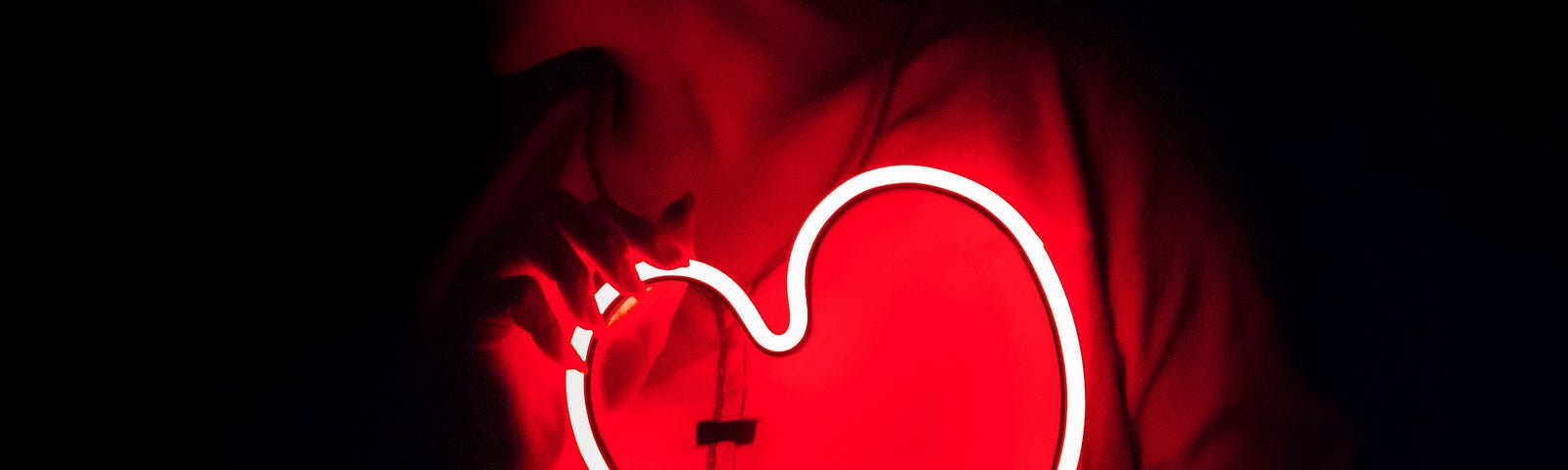 Neon, pink, glowing heart held in front of a woman’s chest against a dark background.
