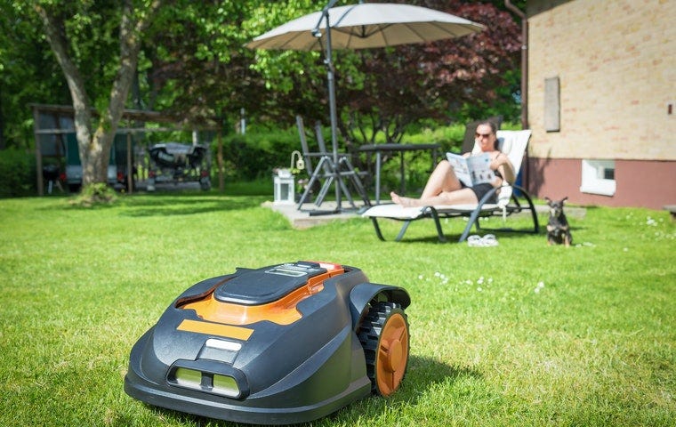 A robotic lawnmower mows the grass while in the background a person reads in a lounge chair.