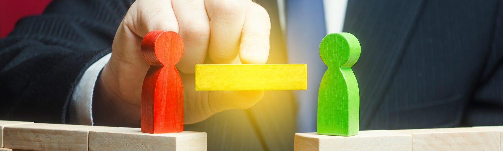 Two block figures stand on two block bridges on a table. A large person wearing a suit behind the table is moving a new block to close the gap between the bridges.