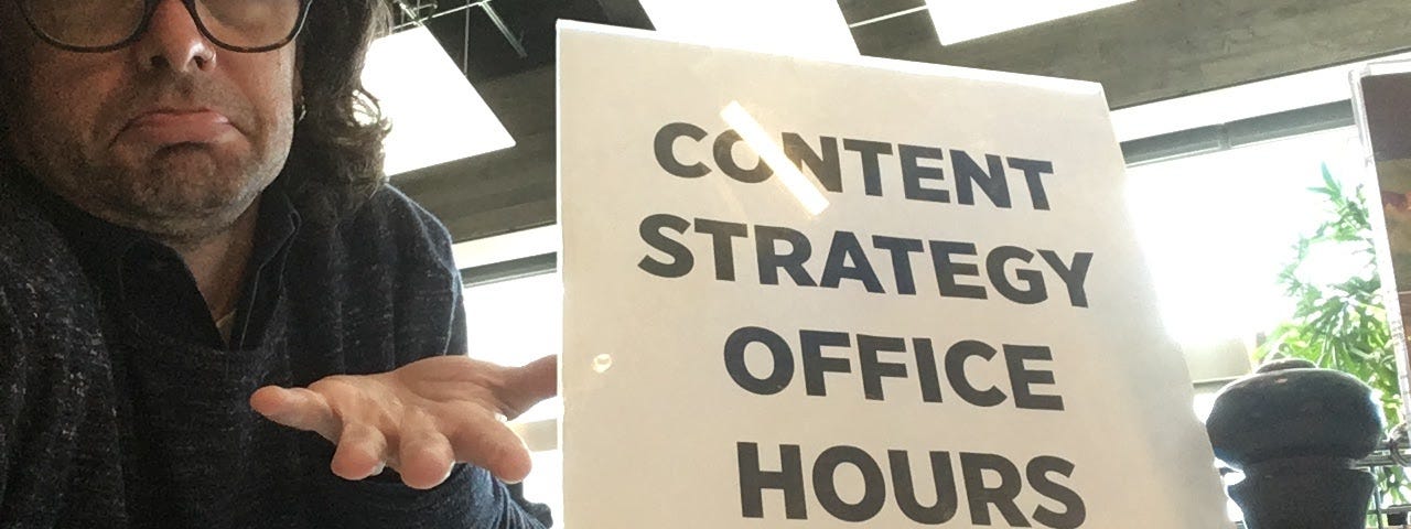 Selfie of me shrugging while sitting at a table next to a sign reading, “Content strategy office hours.”