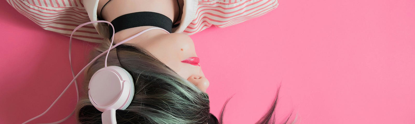Attractive young woman lies down on pink background with pink headphones on. She wears a pink striped blouse and a black ribbon choker. Her hair swirls over her eyes, covering 1/3 of her face.
