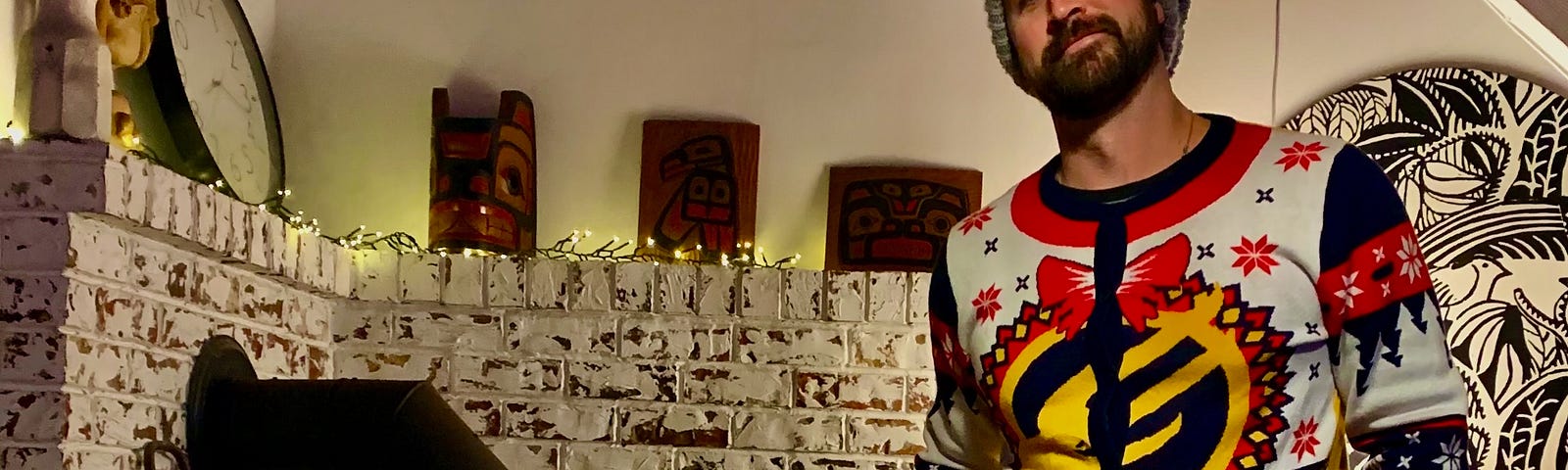 A man stands before a hearth with a wood stove and a brick wall behind him. He is wearing a tuque and an ugly Christmas sweater. He has a beard.