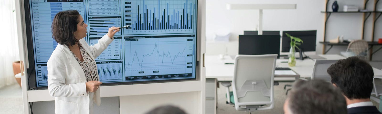 A woman stands in front of a screen displaying multiple graphs and tables, explaining them to a group of men.