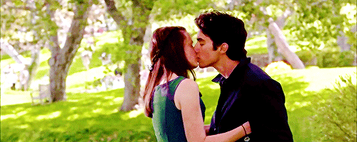 Rory Gilmore, played by Alexis Bledel, is pulling Jess Mariano, played by Milo Ventimiglia, into a surprising kiss. They are outdoors, surrounded by beautiful greenery and sunshine-soaked grass.