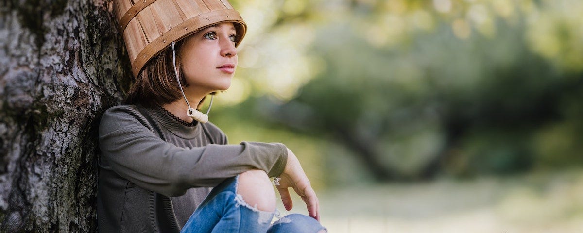 A young girl, perhaps nine years old, sitting against the trunk of a large tree. She has holes in the knees of her jeans, and an upturned wooden bucket on her head, the handle of which hangs like a strap under her chin.