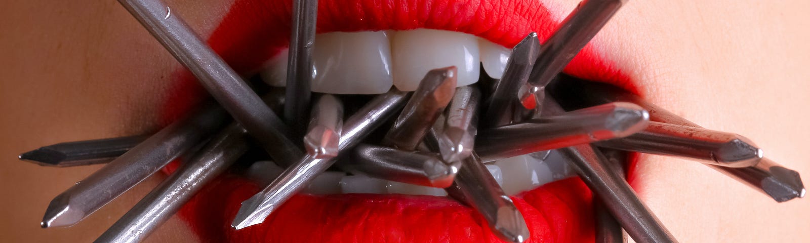 A woman wearing bright red lipstick holds many gray nails in her mouth.
