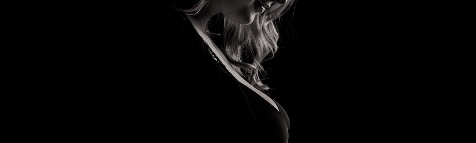 A young woman looks lovingly down as she stands before a black background, her left hand gently placed on her bare, pregnant belly.