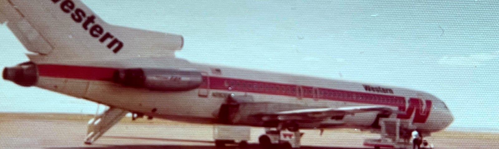 Western Airlines plane on a brown dusty tarmac — white with a red stripe down the side from a large W at the front. Black lettering on the tailfin says “Western”. There is a chain link fence in the foreground with a sign saying “Gates 2–3”