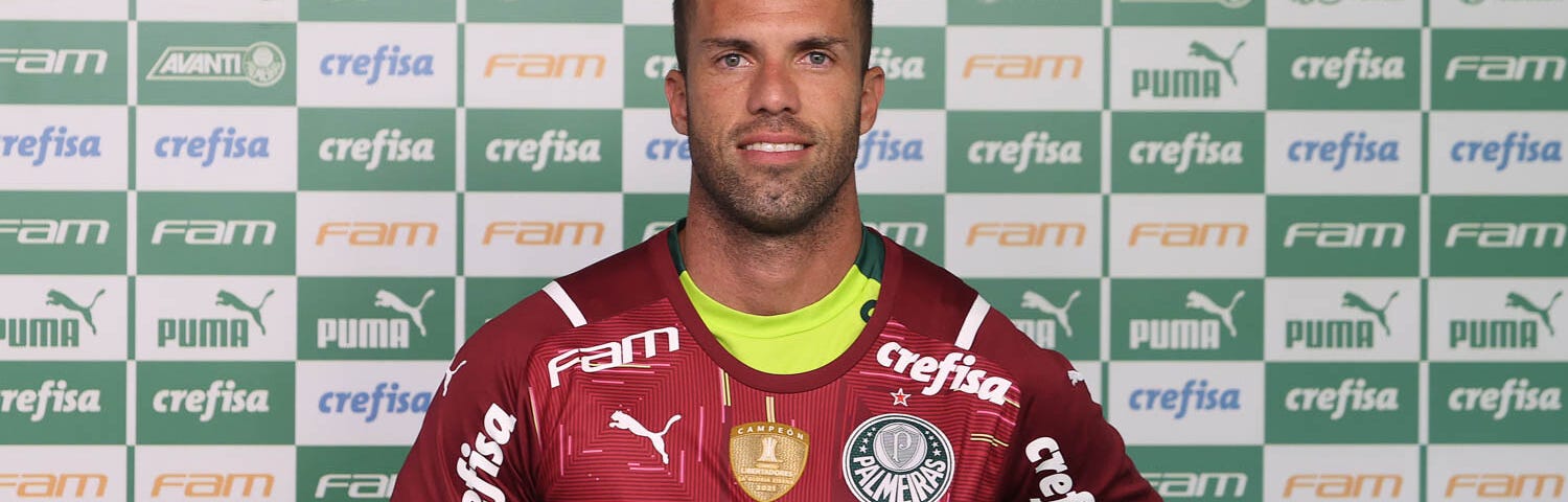 Marcelo Lomba posa com camisa vermelha de goleiro do Palmeiras, à frente de um painel com retângulos verdes e brancos alternando inscrições da Crefisa, da FAM e da Puma. Lomba é um homem branco de pele morena clara, olhos claro, cabeça raspada com máquina 1, barba rala e entradas de calvície.