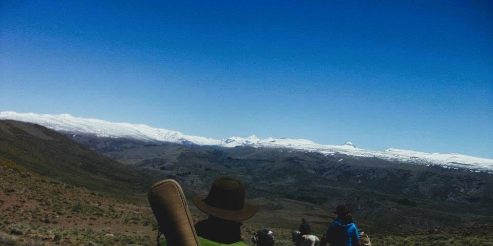 Several people on horseback on a mountain trail headed towards snow capped peaks. One person in the foreground has a guitar on their back.