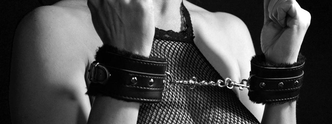 Black & white photo of a woman with plump breasts, a sleeve-less fishnet and with her hands being cuffed by leather handcuffs.