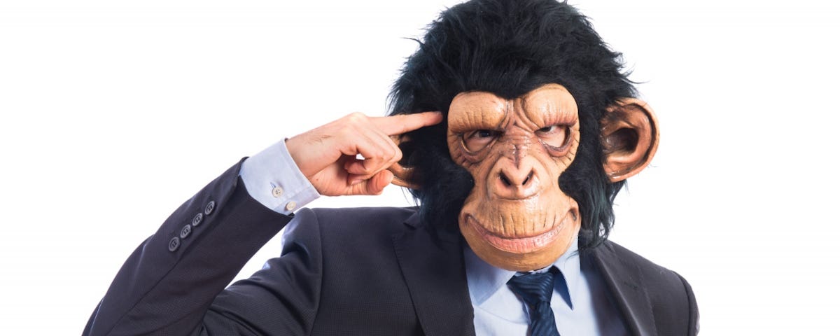 A man in a business suit and a full-head chimp mask pointing at his temple.