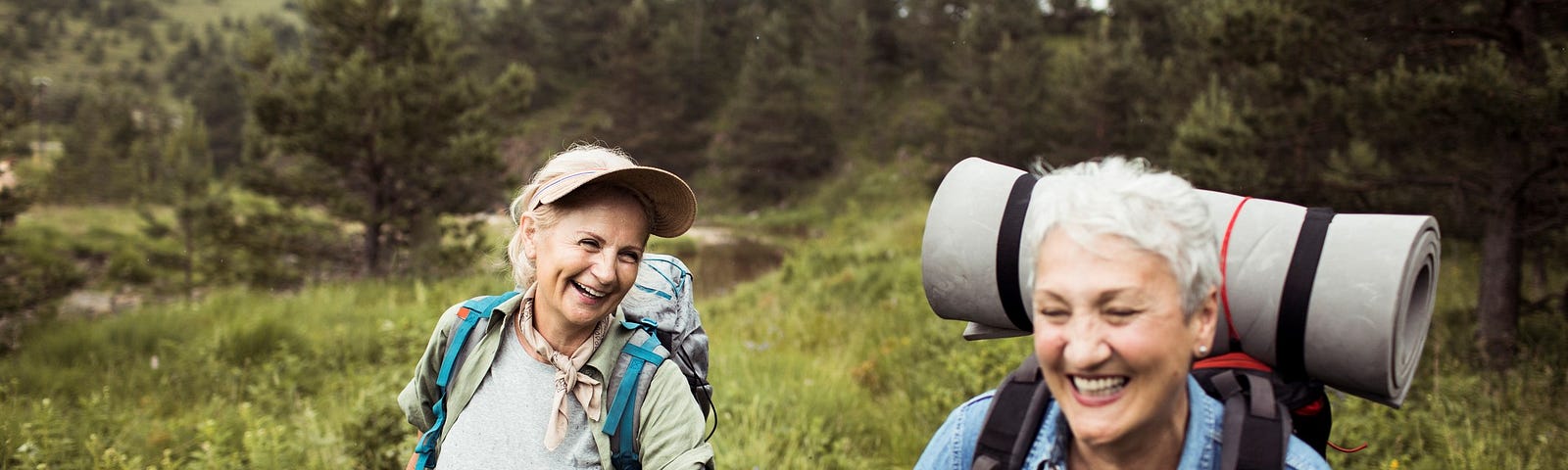 Two older women backpacking through a pale green grassy field, smiling like they’ve just shared a funny inside joke. Dark green pine trees and hills can be seen in the distance.