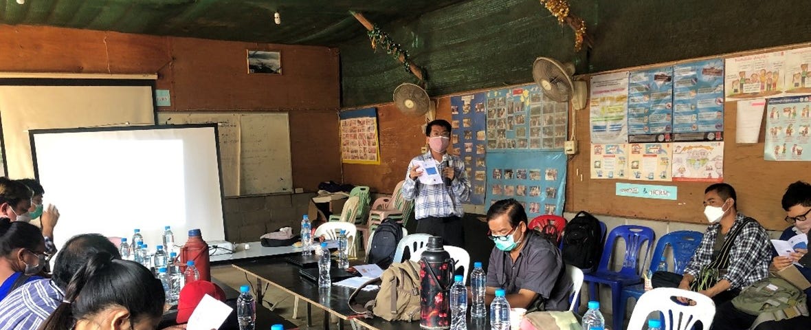 There are around 9 people sitting around a U-shaped table in a room, with one person standing up, facing and speaking to the people sitting around the table. The walls of the room are covered in posters, it looks like a classroom setting.