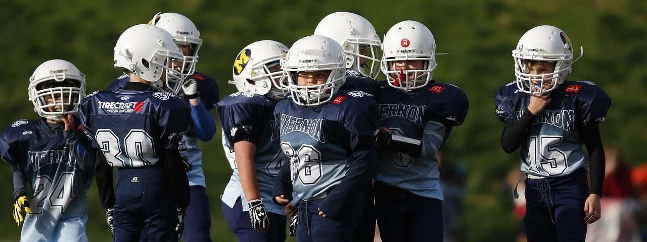 A group of nine maybe six- to eight-year-old football players in full football gear are in a huddle on a turf field ready for their coach to call a play.