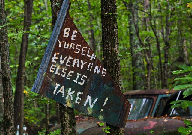 a rusted sign that say “be yourself everyone else is taken”