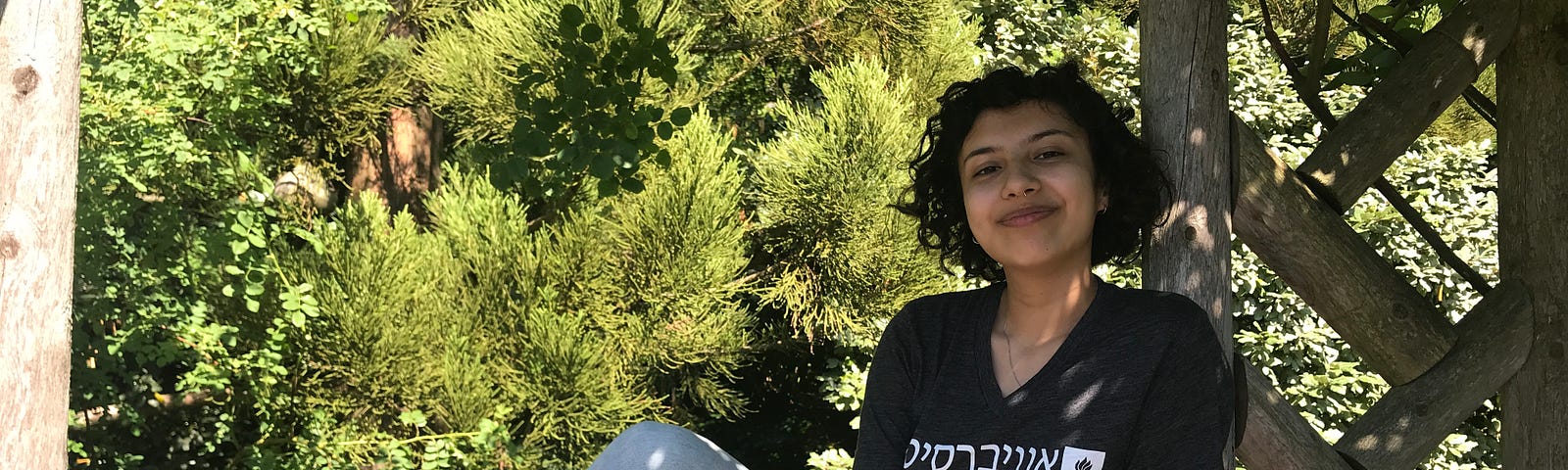 Person wearing black shirt with Hebrew letters and blue jeans sits on wooden horizontal pole with one leg up and one down.