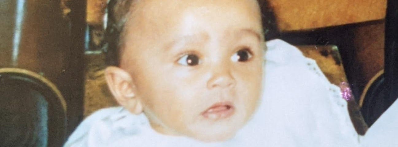 A photograph of me (Kashala, aged around 6 months or so) as a baby, dressed in white, waiting to get christened.