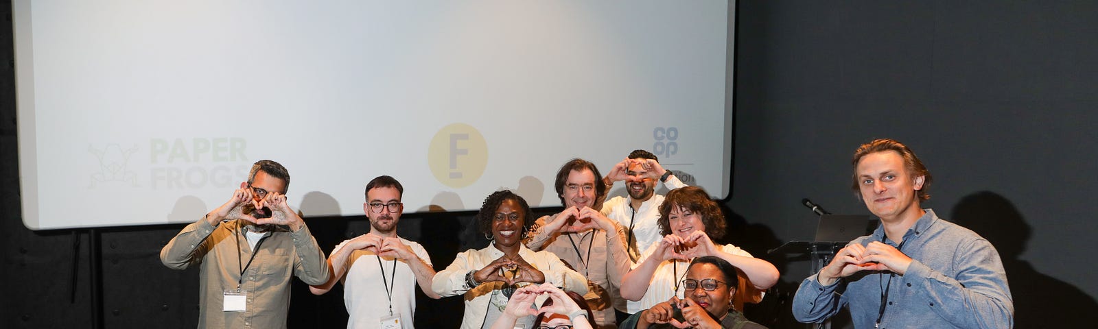 Nine people, making heart shapes with their hands, are grouped in front of a cinema screen that has the words: “Let’s celebrate!”